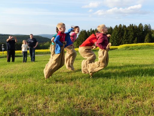 Sackhüpfen Kinder springen in Säcken auf einer Wiese, umgeben von Zuschauern und einer Landschaft.